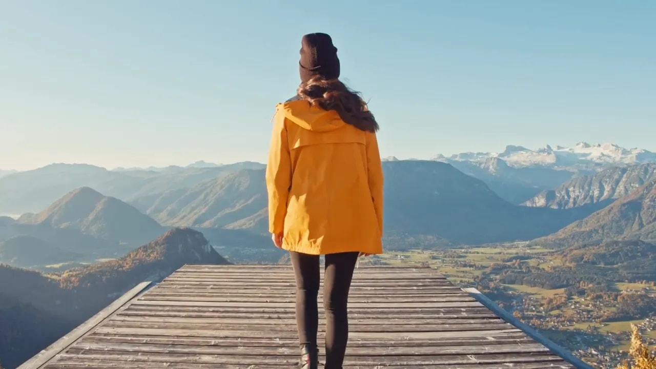 Persoon in geel jack staat op een houten platform en kijkt uit over bergen en een vallei