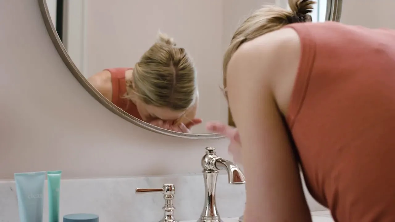 Personne se lavant le visage au lavabo avec un nettoyant et une crème hydratante TrueScience sur le comptoir.